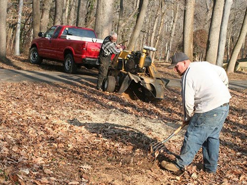 a man in a white shirt raking while another man in overalls prepping a piece of tree removal equipment behind a red pickup truck surrounded by trees
